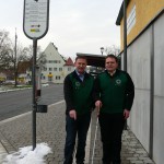 Zwei Männer in grünen Westen stehen an einer Bushaltestelle mit Schnee am Boden.
