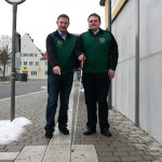 Zwei Männer in grünen Westen stehen neben einem Bordstein auf einem Gehweg.