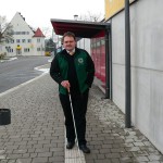 Mann mit Blindenstock steht auf dem Gehweg eines modernen Gebäudes.