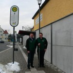 Zwei Männer stehen an einer Haltestelle neben einem grauen Gebäude. Schnee liegt auf dem Boden.