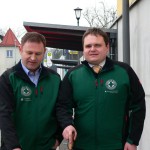 Zwei Männer in grünen Jacken gehen nebeneinander auf einer Straße.